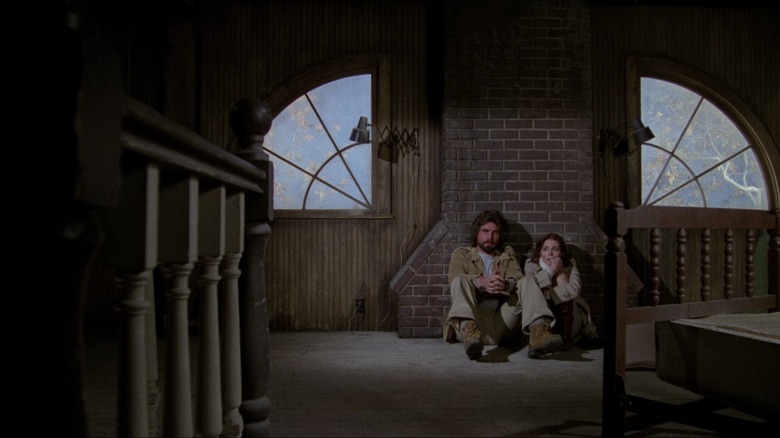 James Brolin as George Lutz and Margot Kidder as Kathy Lutz in The Amityville Horror, sitting on the ground next to each other against a brick wall in a dark, creepy-looking part of the house.