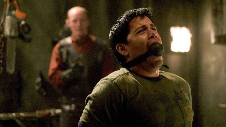 Jay Hernandez as Paxton sitting with his arms tied behind his back and a ball gag in his mouth looking distressed as a man stands behind him with a rubbed apron on in Hostel