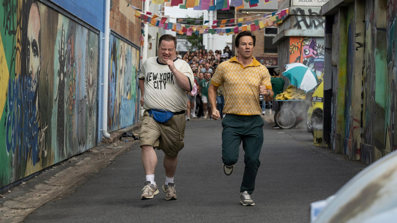 Mark Wahlberg as Brad Lewison and Paul Walter Hauser as Elijah DeBell in Balls Up, running down an alleyway with a crowd behind them.