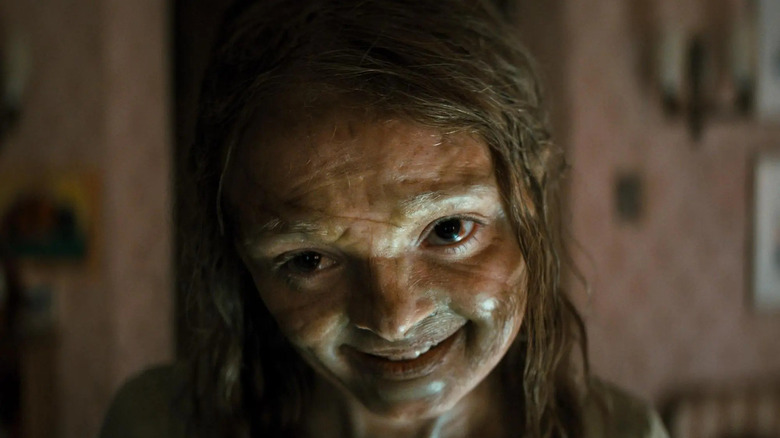 Natalie Grace as Katie Cannon in Lee Cronin's The Mummy, looking dirty and slightly deformed while she creepily smiles.