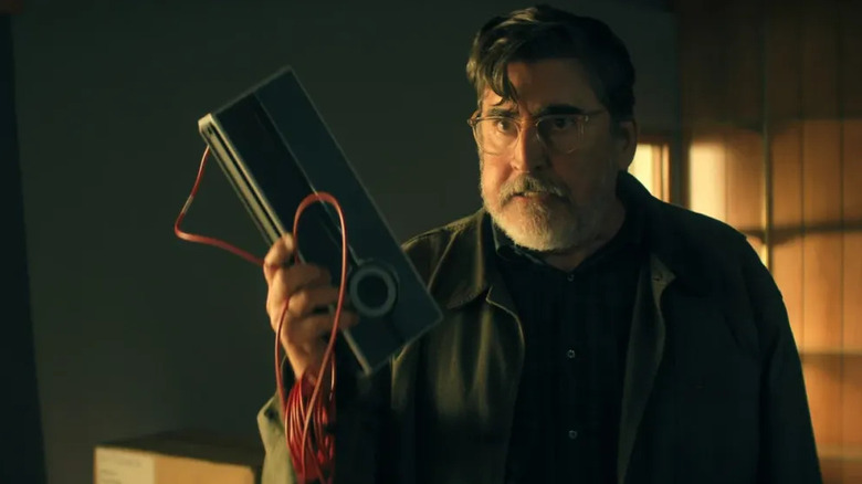 Alfred Molina as Sam Cooper in The Boroughs, holding up an electronic device and looking disgruntled.
