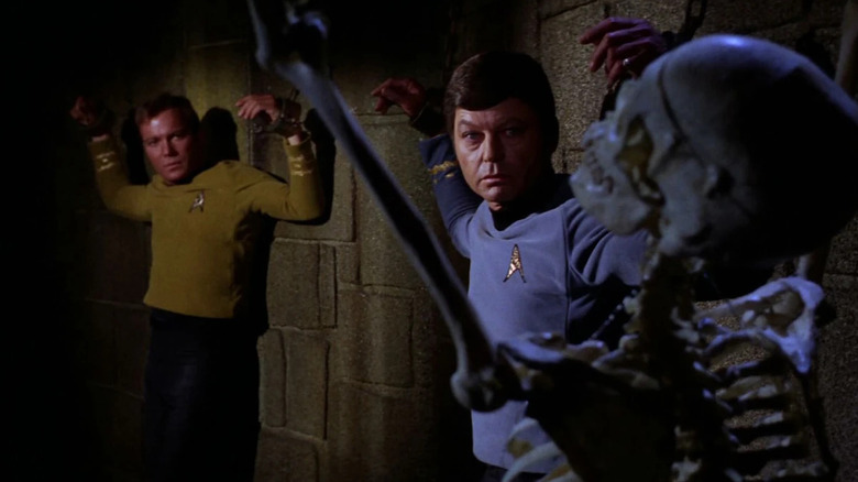 Kirk (William Shatner) and Leonard McCoy (DeForest Kelley) glance at a skeleton while chained to a wall in Star Trek: The Original Series
