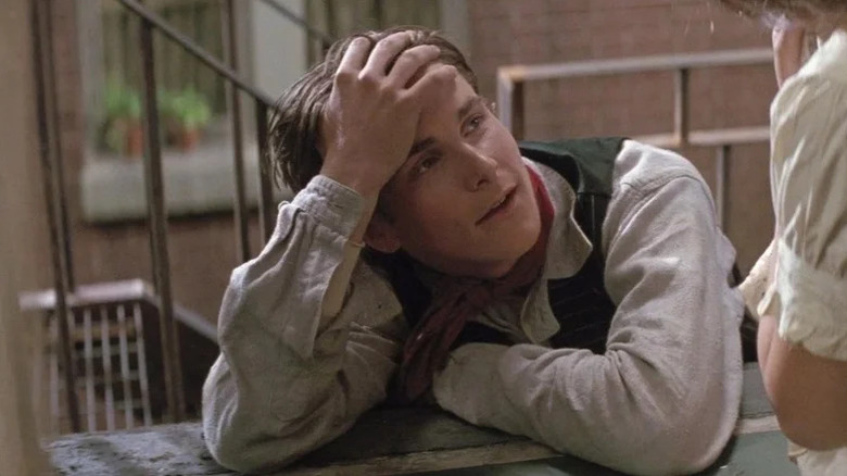 Christian Bale in vest, white shirt and red ascot as Cowboy leaning with hand on his head in Newsies