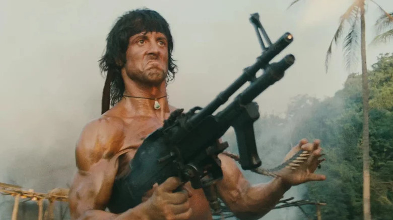 Sylvester Stallone as John Rambo shirtless in jungle environment holding machine gun and grimacing in Rambo: First Blood Part II