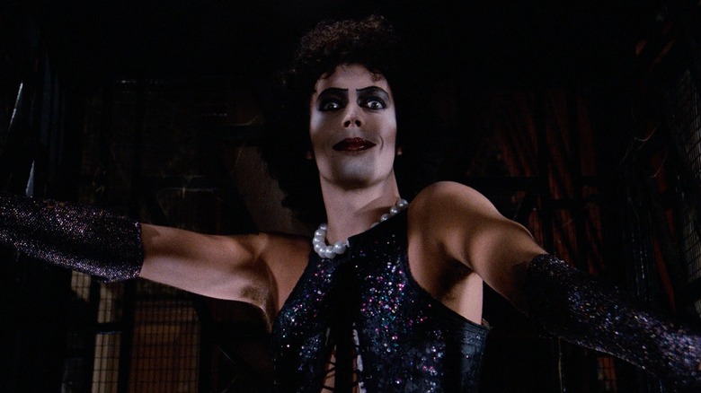 Tim Curry as Dr. Frank-N-Furter in The Rocky Horror Picture Show, wearing a black sequined halter corset and gloves as he extends his arms and looks toward the camera.