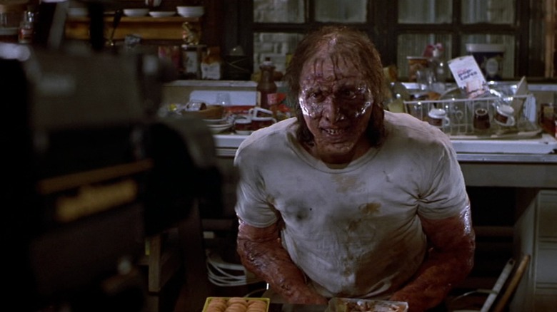 Jeff Goldblum as Seth Brundle in The Fly, face and arms covered in gloppy boils, sitting in front of a camera