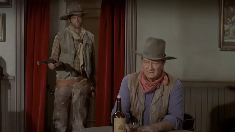 McNally (John Wayne) looks at a holdup while sitting with a glass of whiskey and a henchman behind him in Rio Lobo