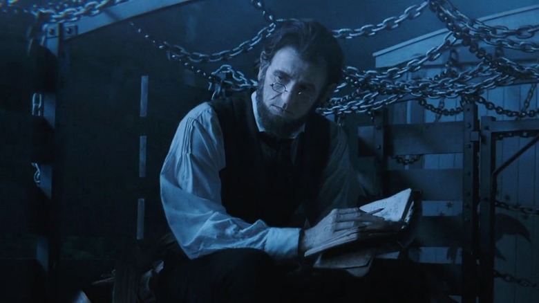 Benjamin Walker as Abraham Lincoln in Abraham Lincoln: Vampire Hunter, sitting in a dark blue environment among chains, bespectacled and with a journal open in his hands.