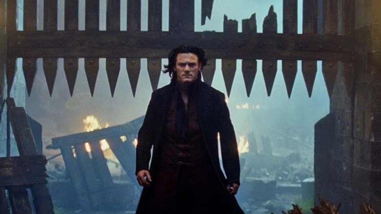 Luke Evans as Vlad III "the Impaler" Dracula in Dracula Untold, walking away from a half-open castle gate, with fire and destruction behind him.