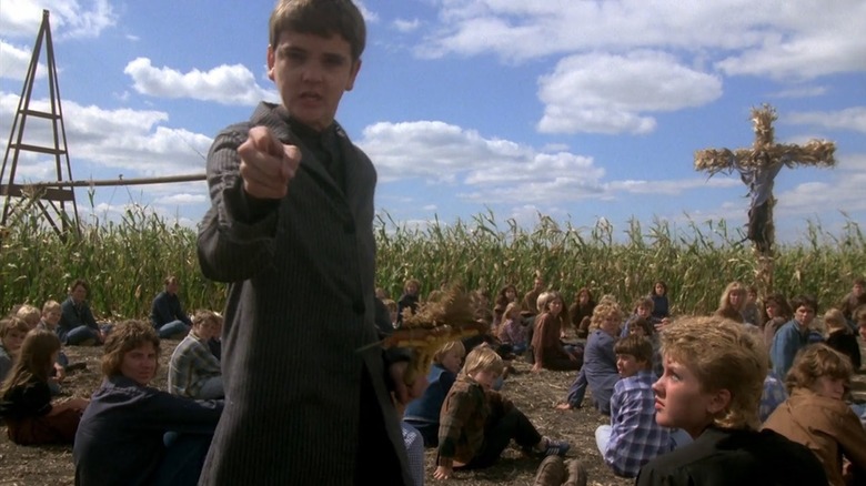 John Franklin as Isaac in Children of the Corn, pointing toward the camera with a cabal of other children sitting around behind him in the daylight, all in front of a cross made out of crops.