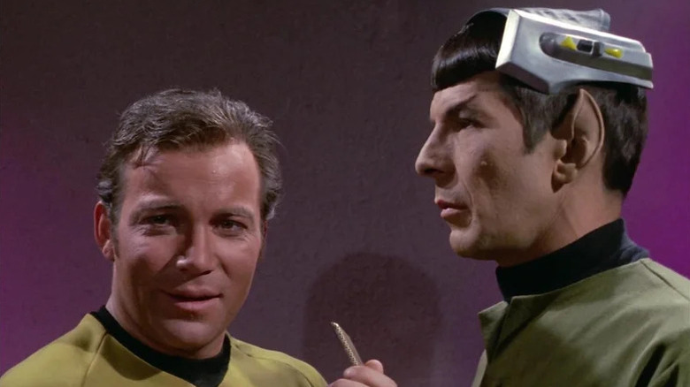 A bemused Jim Kirk (William Shatner) speaks on a communicator next to Spock (Leonard Nimoy) wearing a cranial device in Star Trek: The Original Series