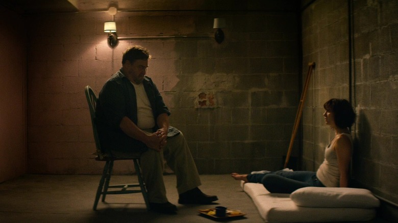 Howard sitting on a chair with Michelle on a mattress in 10 Cloverfield Lane