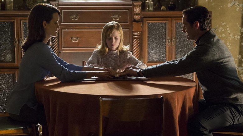 Elizabeth Reaser as Alice Zander, Lulu Wilson as Doris Zander, and Henry Thomas as Father Thomas Hogan seated around a ouija board in Ouija: Origin of Evil