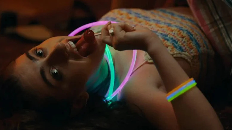 Rachel Sennott as Alice laying down, wearing glowsitcks and licking lollipop in Bodies Bodies Bodies