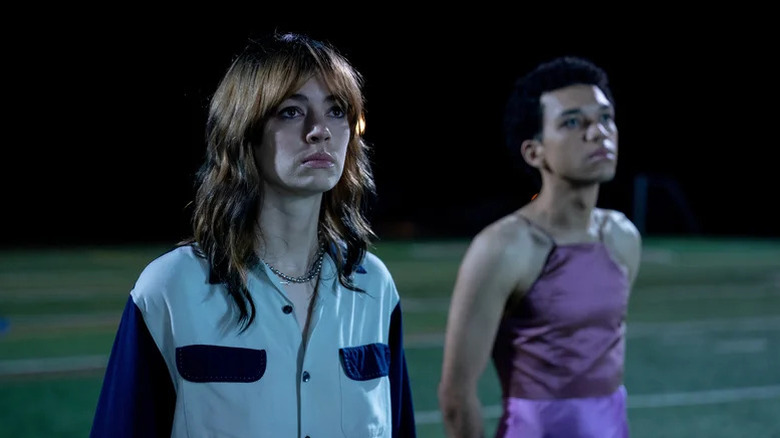 Jack Haven as Maddy in blue jacket standing next to Justice Smith as Owen in pink dress on green sports field in I Saw the TV Glow