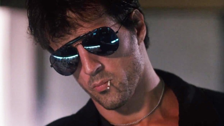 Marion Cobretti (Sylvester Stallone) looks down, wearing big sunglasses with a match in his mouth in Cobra (1986)