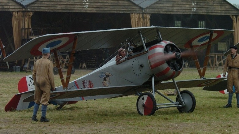 Two mechanics gather around a pilot and a biplane by some hangars in Flyboys