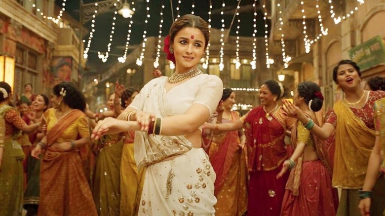 Alia Bhatt as Gangubai wearing white sari in the street in front of women wearing red and orange saris in Gangubai Kathiawadi