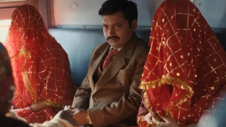 Bhaskar Jha as Pradeep Singh, wearing a mustache and brown suit sitting next to two veiled women in red on a train in Lost Ladies