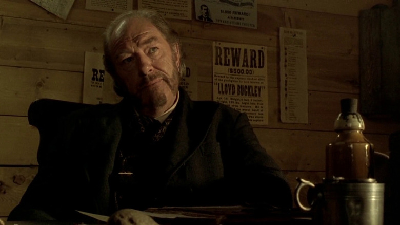 Michael Gambon as Denton Baxter sitting at a desk in front of wanted posters in Open Range