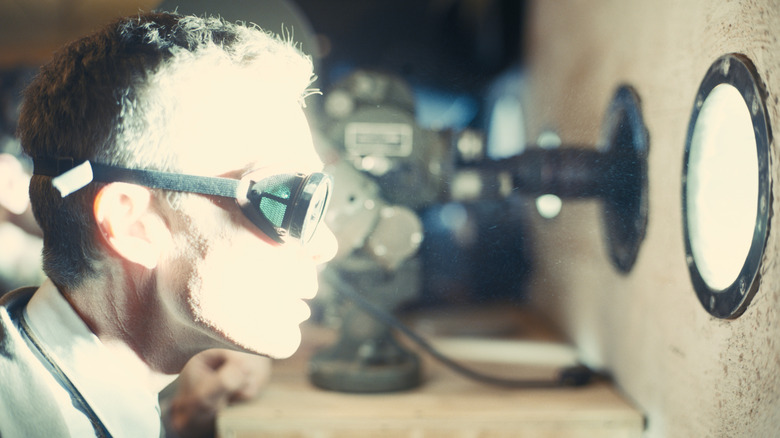 Cillian Murphy as J. Robert Oppenheimer wearing goggles and watching a distant explosion in Oppenheimer