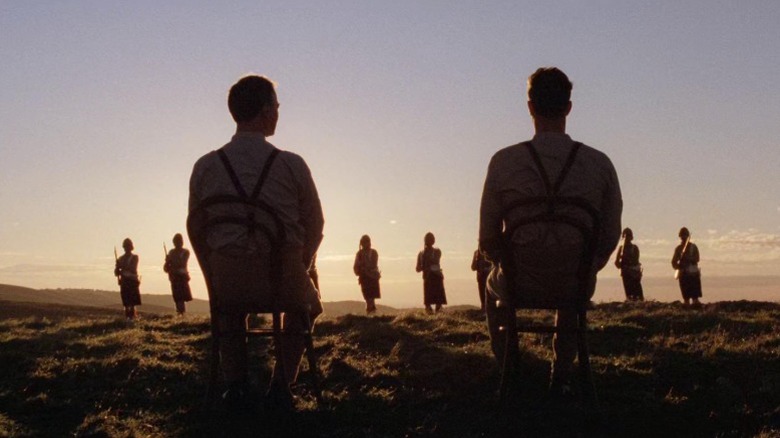 Edward Woodward as Harry "Breaker" Morant and Bryan Brown as Peter Handcock sitting on chairs before a row of armed soldiers in a field in Breaker Morant
