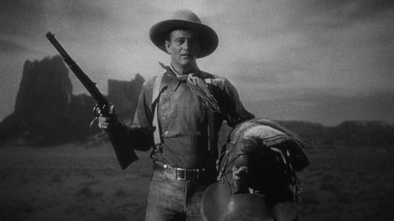 John Wayne as Henry, the Ringo Kid, holding a gun and a saddle in the desert in Stagecoach