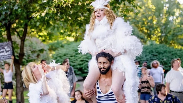 Natasha Lyonne sitting on man's shoulders in white boa outfit