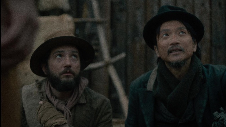 John Magaro as Cookie and Orion Lee as King-Lu in First Cow, sitting side by side and looking up at somebody.