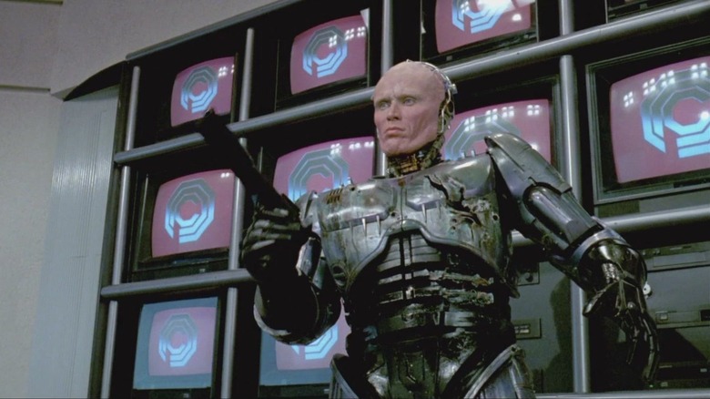 Peter Weller as Alex Murphy / RoboCop in RoboCop, face exposed and wielding his weapon in front of a grouping of televisions.