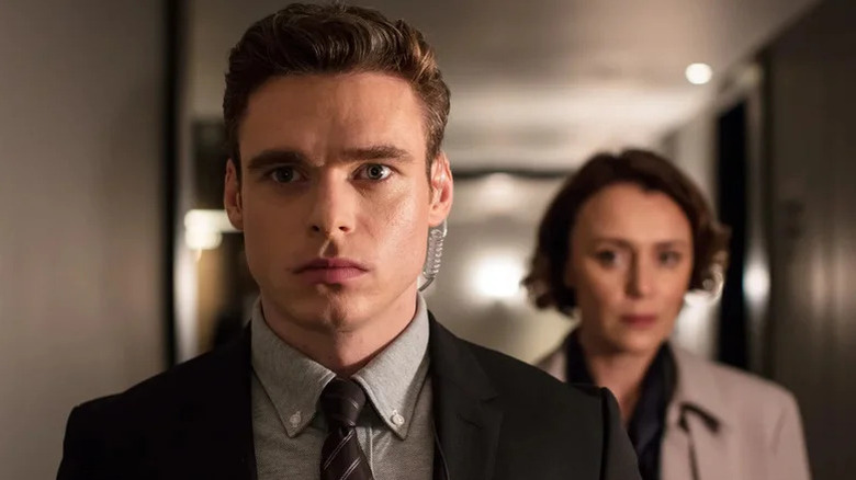 Richard Madden David Budd in suit with earpiece in standing in front of Keeley Hawes as Julia Montague (not in focus) in Bodyguard