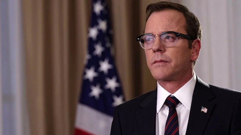 Kiefer Sutherland as Thomas Kirkman in suit and glasses standing in front of American flag