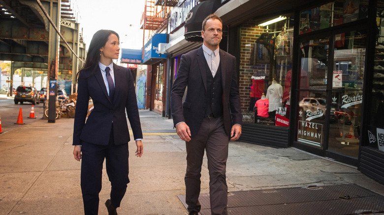 Joan Watson (Lucy Liu) and Sherlock Holmes (Jonny Lee Miller) walk down a Brooklyn sidewalk wearing suits on Elementary