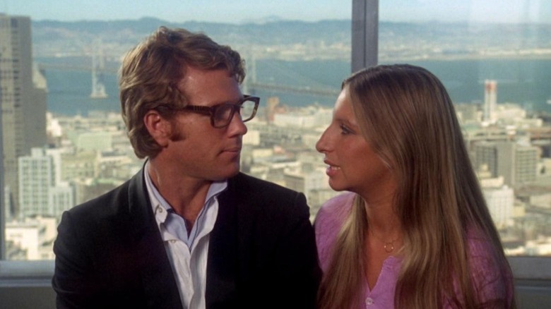 Ryan O'Neal as Howard Bannister and Barbra Streisand as Judy Maxwell sitting side by side in front of a skyscraper window in What's Up, Doc?