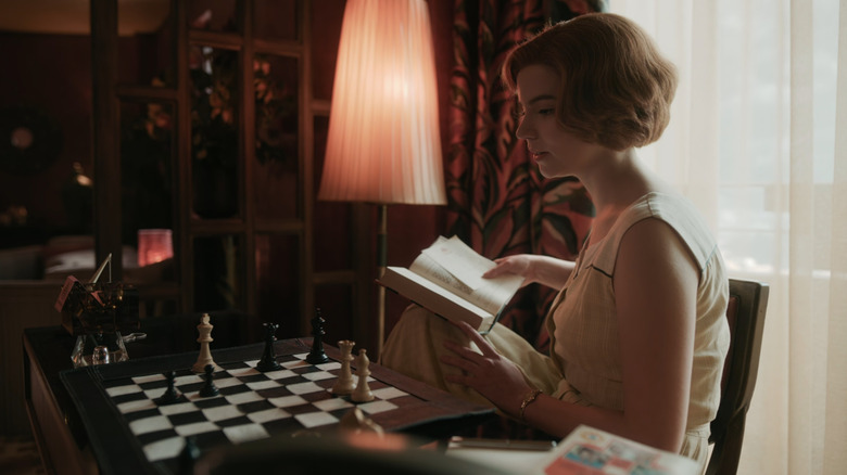 Beth (Anya Taylor-Joy) holding book and staring at chess board in The Queen's Gambit