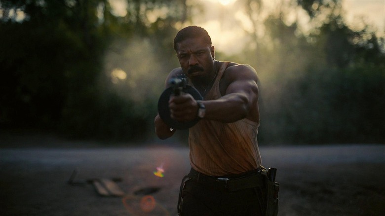 Michael B. Jordan as Smoke in Sinners, pointing a Tommy gun right past the camera against a rising sun.