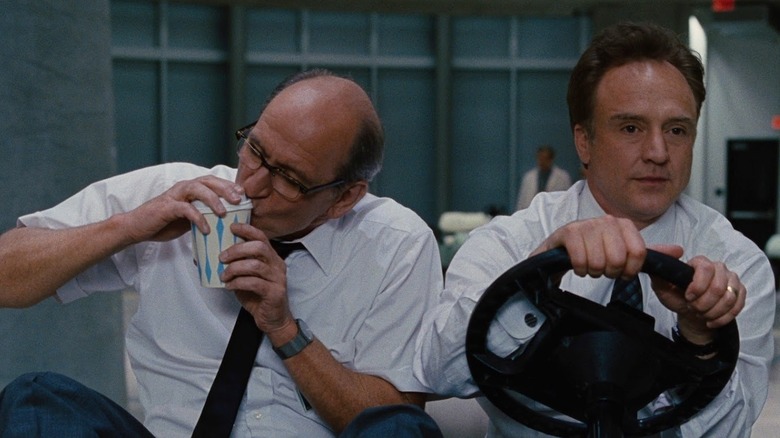 Richard Jenkins as Gary Sitterson and Bradley Whitford as Steve Hadley in The Cabin in the Woods, with Gary trying to sip out of a coffee cup while Steve drives them on a golf cart.
