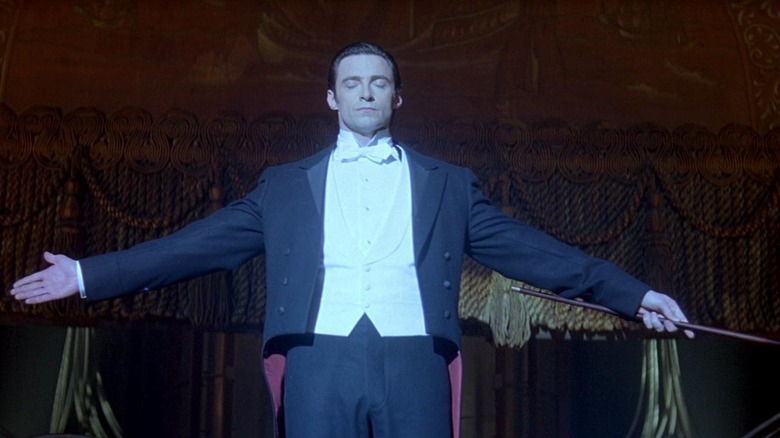 Hugh Jackman as Robert "The Great Danton" Angier / Lord Caldlow in The Prestige, spotlighted on stage with his arms spread open and eyes closed.