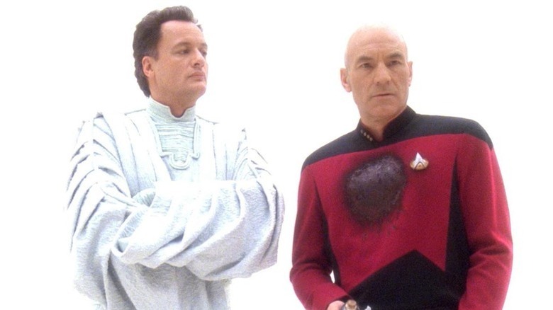 Q (John de Lancie) stands solemnly next to Jean-Luc Picard (Patrick Stewart) with a bloody chest in a bright white space in Star Trek: The Next Generation