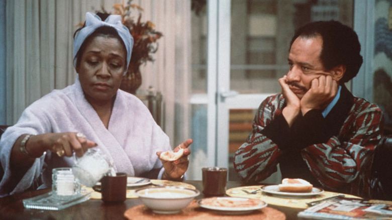 Isabel Sanford as Louise "Weezy" Jefferson sits at a table next to Sherman Hemsley as George Jefferson in The Jeffersons.
