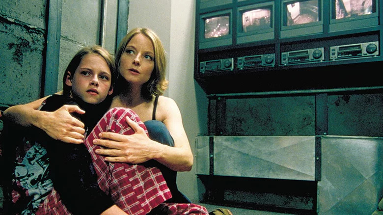 Young Kristen Stewart as Sarah sitting and Jodie Foster as Meg with arms around her in cement room with security footage behind them in Panic Room