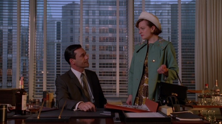 Jon Hamm as Don Draper sitting at his desk while Elisabeth Moss as Peggy Olson stands next to him on Mad Men
