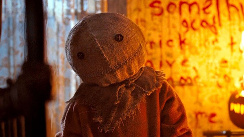 Child in red pajamas with cloth pumpkin over his head (Quinn Lord) cocking his head in front of wall with bloody writing