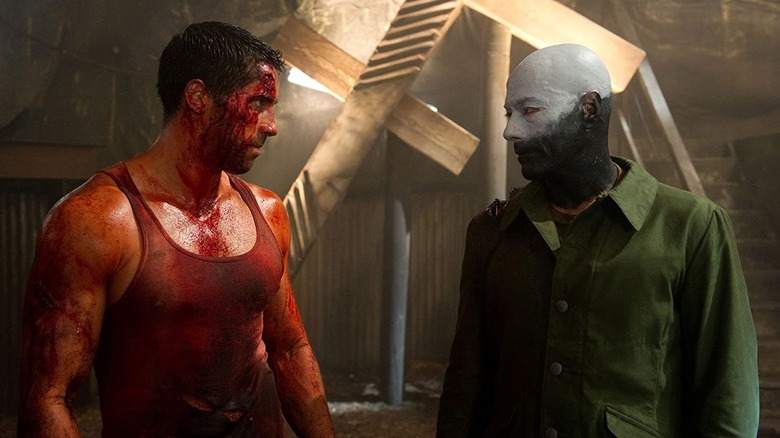 John (Scott Adkins) in tank top covered in blood standing next to Luc Devereaux (Jean-Claude Van Damme) in green jacket with top of his face painted white and the bottom painted black