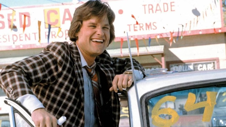 Rudy Russo (Kurt Russell) smiles leaning on a used car for sale in Used Cars