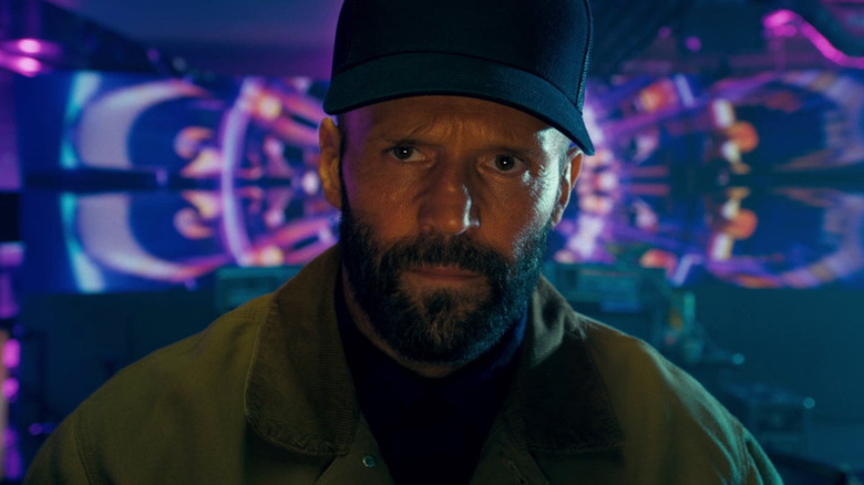Jason Statham as Adam Clay stands in front of neon lights in The Beekeeper