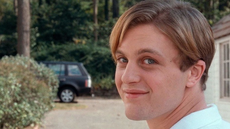 Michael Pitt as Paul smiling at the camera with a car and house in the background in Funny Games