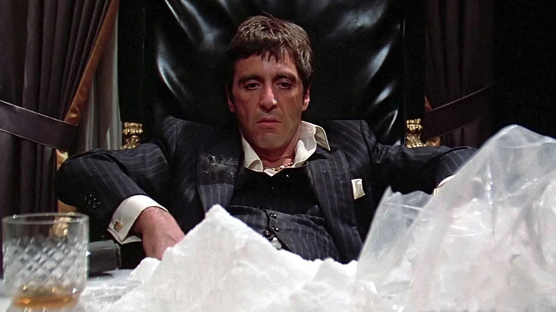 Al Pacino sitting at a desk in a black chair with a drink and drugs in front of him in Scarface