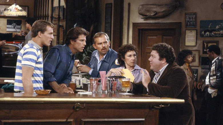 Woody Harrelson, Ted Danson, John Ratzenberger, Rhea Perlman, and George Wendt as Woody Boyd, Sam Malone, Cliff Clavin, Carla Tortelli, and Norm Peterson gathering around Norm's stool at the bar in Cheers