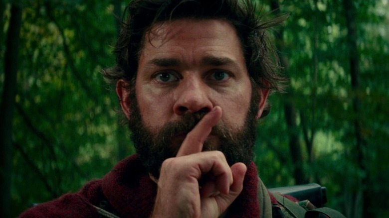 A close-up of John Krasinski as Lee Abbott in A Quiet Place, standing within a forest and holding his finger up to his mouth in a "be quiet" position.